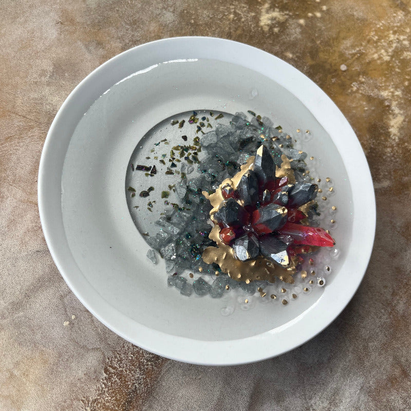 A dark pewter colored epoxy crystal cluster with pink accents, floating over a gray crushed rock lake, with painted gold accents, presented on a white ceramic tray. Overhead View