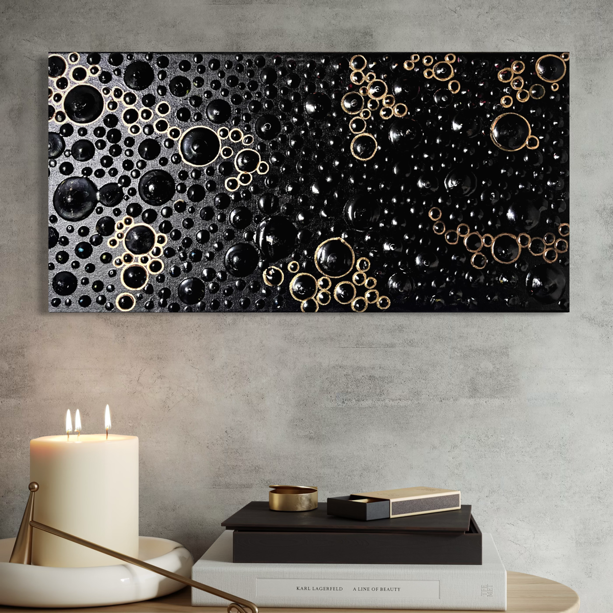 A rectangular black abstract wall art piece with raised circular bubble-like forms in black and metallic gold, displayed on a light gray wall above a styled tabletop with a candle, books, and small decor objects.