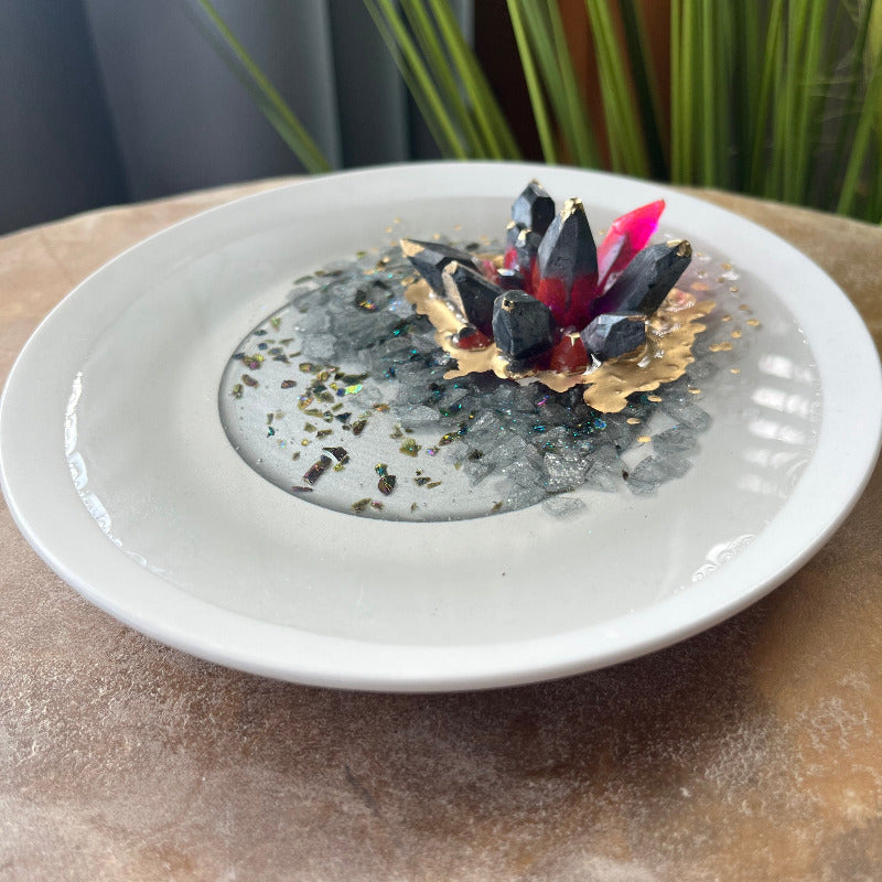 A dark pewter colored epoxy crystal cluster with pink accents, floating over a gray crushed rock lake, with painted gold accents, presented on a white ceramic tray. Angled View