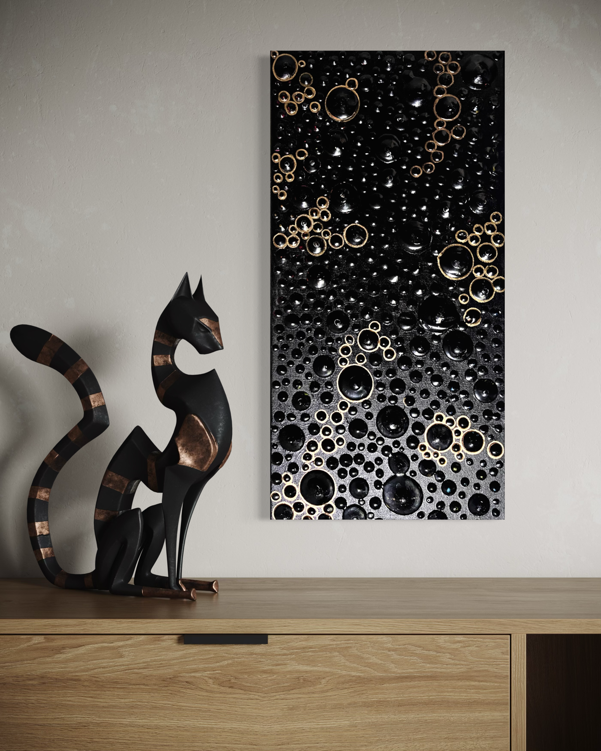 A vertical black canvas wall art piece with a gradient of metallic gold and silver bubble-like rings, displayed on a light gray wall above a wooden console table next to a black cat sculpture.