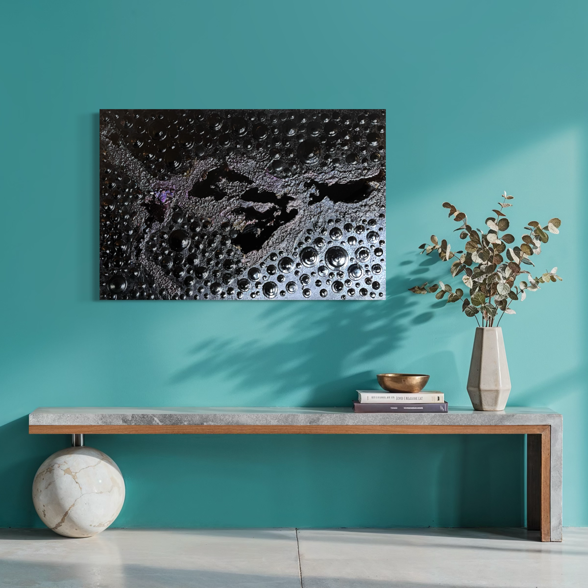 A square abstract wall painting with a black bubbly texture  and smooth glossy lakes hangs on a teal wall above a modern console table with a vase of eucalyptus and books.