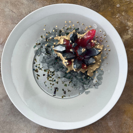 A dark pewter colored epoxy crystal cluster with pink accents, floating over a gray crushed rock lake, with painted gold accents, presented on a white ceramic tray. Overhead View