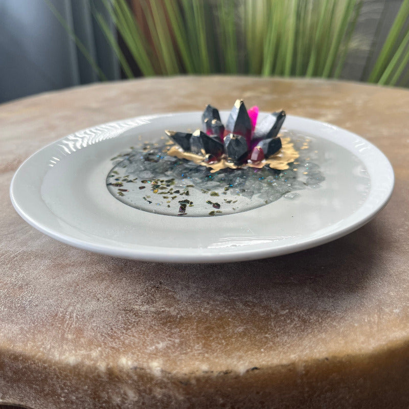 A dark pewter colored epoxy crystal cluster with pink accents, floating over a gray crushed rock lake, with painted gold accents, presented on a white ceramic tray. angled side View