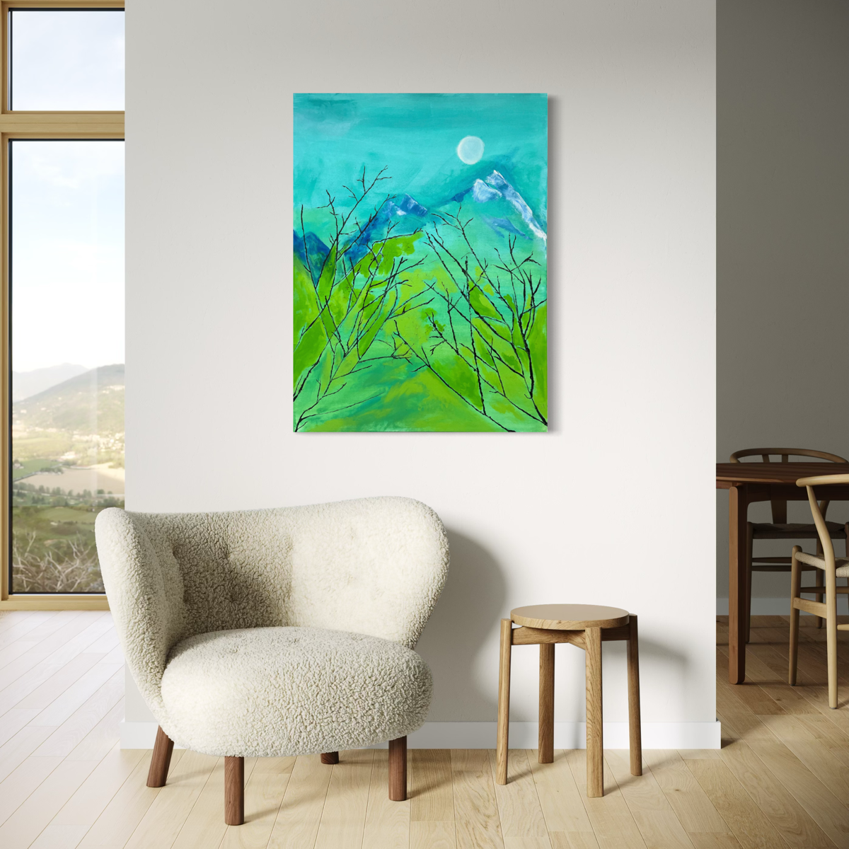 A vertical canvas art print depicting a vibrant green landscape with blue mountains and a white moon, displayed on a white wall in a modern living room setting with a cream boucle armchair and a small wooden stool.